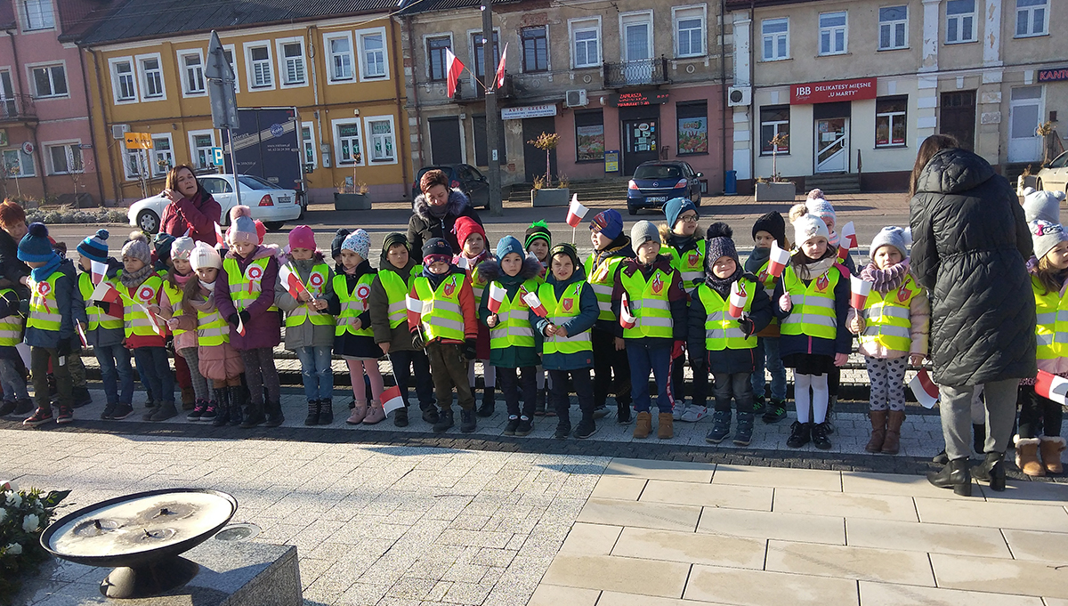 Dzieci pod pomnikiem w parku miejskim, trzymają flagi Polski