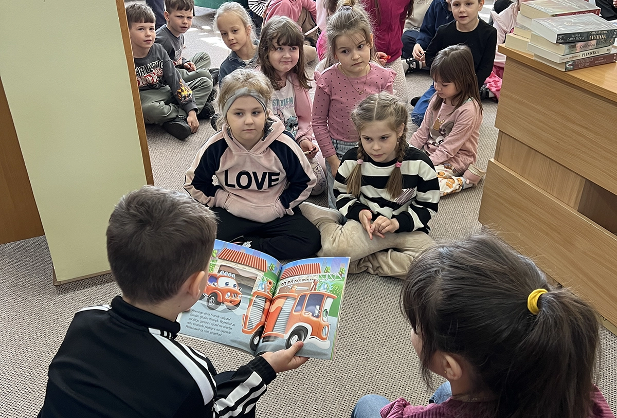 dzieci słuchają jak chłopiec czyta książkę w bibliotece